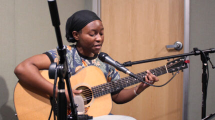 An adult playing acoustic guitar and singing into a microphone in a radio studio
