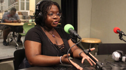A teen wearing headphones and speaking into a microphone gesturing with their hands in a radio studio