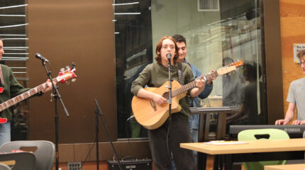 An adult playing electric bass, an adult playing acoustic guitar and singing into a microphone, an adult playing acoustic guitar, and an adult playing a piano keyboard