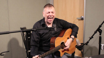 An adult playing acoustic guitar and singing into a microphone in a radio studio