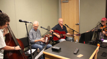 An adult playing upright bass, an adult playing accordion, an adult playing acoustic guitar, and an adult playing fiddle in a radio studio
