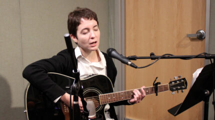 An adult playing acoustic guitar and singing into a microphone sitting behind sheet music on a music stand in a radio studio