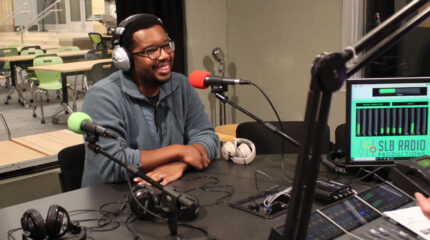 An adult wearing headphones smiling behind a microphone being interviewed in a radio studio