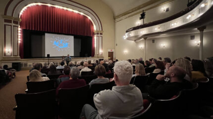 An audience looking at a screen with a red curtain above in an auditorium