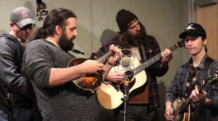 An adult playing upright bass, an adult playing mandolin, an adult playing acoustic guitar, and an adult playing banjo, all standing around a microphone in a radio studio