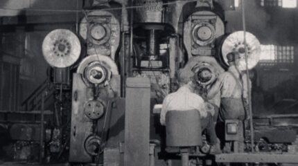 Black and white image of two adults in front of a large machine in a factory