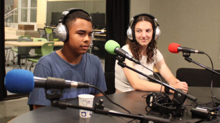 Two teens wearing headphones and sitting behind microphones in a radio studio
