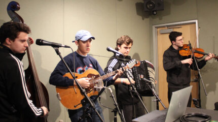 An adult playing upright bass, an adult playing acoustic guitar, an adult playing accordion, and an adult playing viola, all standing behind microphones in a radio studio