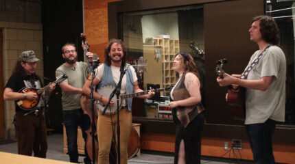 An adult playing mandolin, an adult playing upright bass, an adult playing banjo, an adult singing into a microphone, and an adult playing acoustic guitar