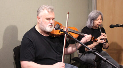 An adult playing a fiddle into a microphone and an adult playing a whistle into a microphone in a radio studio