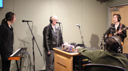 Three adults wearing earphones and standing behind microphones, one holding an acoustic guitar, one standing behind a synthesizer, as another adult wearing headphones listens in a radio studio