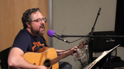 An adult playing acoustic guitar and singing into a microphone looking at sheet music on a music stand in a radio studio