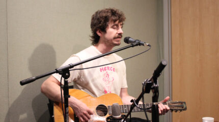 An adult playing acoustic guitar and singing into a microphone in a radio studio