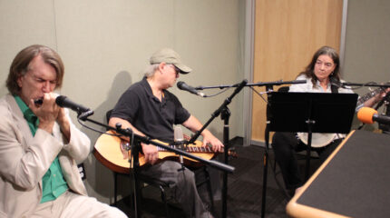 An adult playing harmonica into a microphone, an adult playing dulcimer, and an adult playing banjo looking at sheet music on a music stand in a radio studio