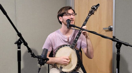 An adult playing banjo and singing into a microphone in a radio studio