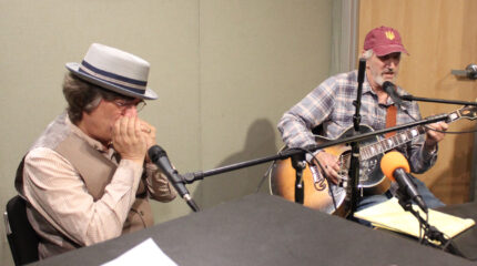 An adult playing harmonica into a microphone and an adult playing acoustic guitar and singing into a microphone in a radio studio