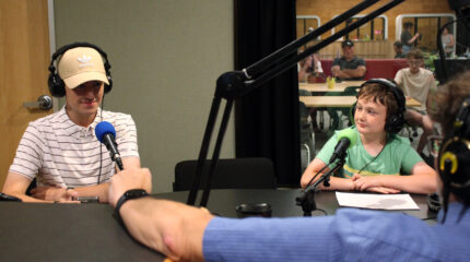 Two youth wearing headphones sitting behind microphones being interviewed by an adult wearing headphones in a radio studio