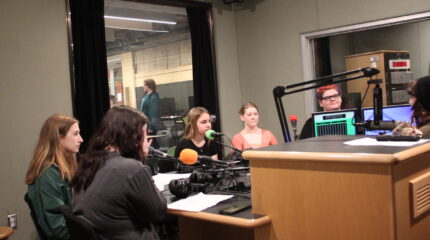 Five teens sitting behind microphones being interviewed by an adult sitting behind a microphone in a radio studio