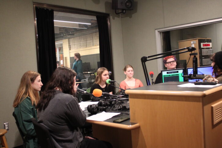 Five teens sitting behind microphones being interviewed by an adult sitting behind a microphone in a radio studio