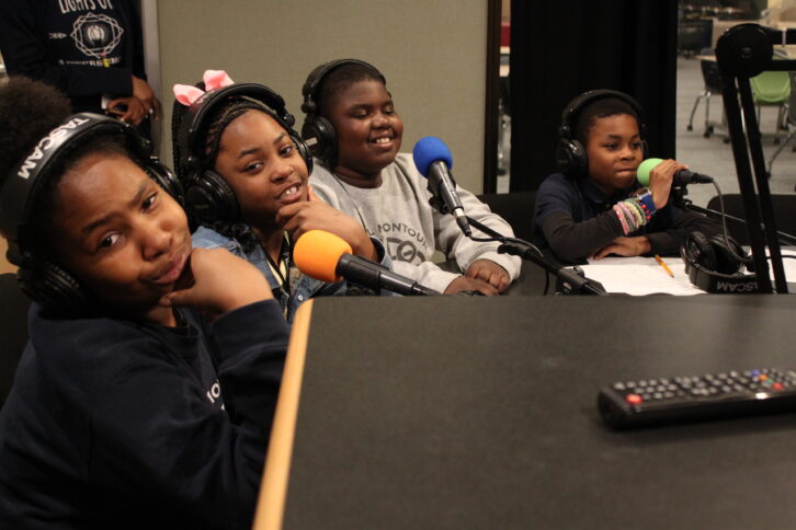 Four youth wearing headphones and smiling sitting behind microphones in a radio studio