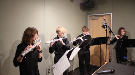 Two teens playing flutes, a teen playing alto flute, and a teen playing bass flute, all looking at sheet music on music stands in a radio studio