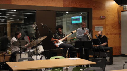 An adult playing a drum set, an adult playing an electric bass, an adult playing a soprano saxophone into a microphone, and an adult wearing a mask playing a piano keyboard