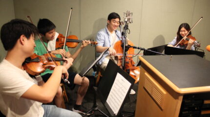 Two teens playing violin, a teen playing cello, and a teen playing viola, all looking at sheet music on music stands in a radio studio