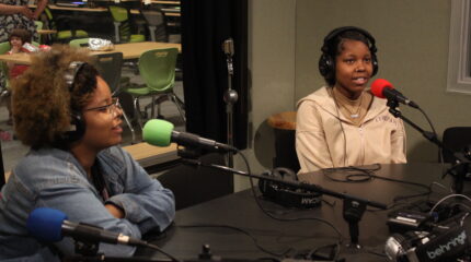 Two teens wearing headphones speaking into microphones in a radio studio