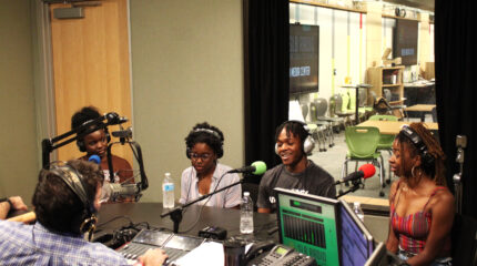 Four teens wearing headphones speaking into microphones being interviewed by an adult wearing headphones sitting behind a microphone and a mixing board in a radio studio