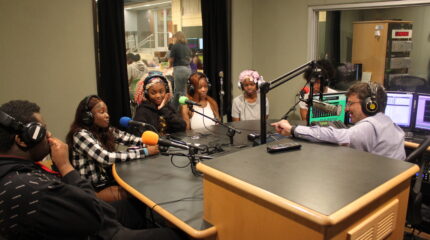 Five teens wearing headphones speaking into microphones being interviewed by an adult wearing headphones sitting behind a microphone in a radio studio