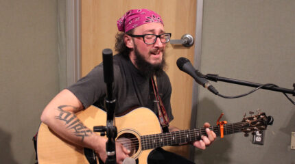 An adult playing acoustic guitar and singing into a microphone in a radio studio