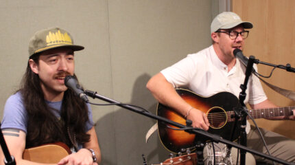 Two adults singing into microphones and playing acoustic guitars in a radio studio
