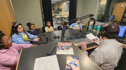 One adult and five youth, all wearing headphones and sitting behind microphones, being interviewed by an adult wearing headphones sitting behind a microphone and a mixing board in a radio studio