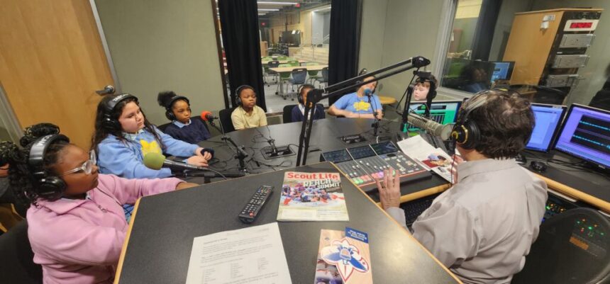 One adult and five youth, all wearing headphones and sitting behind microphones, being interviewed by an adult wearing headphones sitting behind a microphone and a mixing board in a radio studio