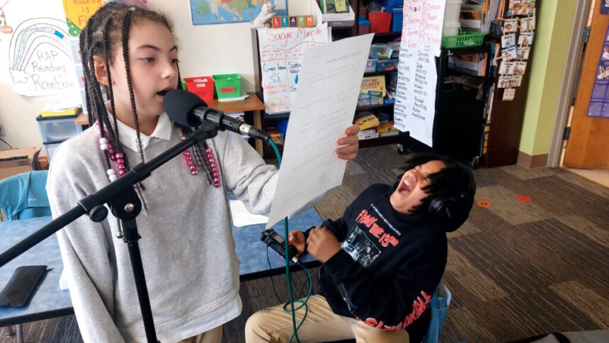 A youth wearing headphones reading from a piece of paper into a microphone as another youth poses in a scream kneeling on the ground