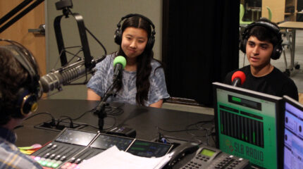Two teens wearing headphones speaking into microphones being interviewed by an adult wearing headphones sitting behind a microphone and a mixing board in a radio studio