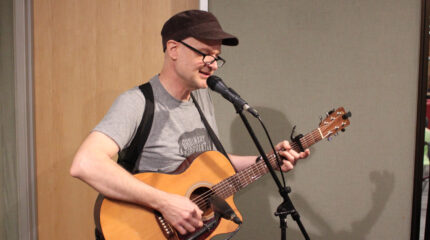 An adult playing acoustic guitar and singing into a microphone in a radio studio