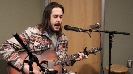 An adult playing acoustic guitar and singing into a microphone in a radio studio