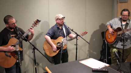 Three adults playing acoustic guitars and singing into microphones in a radio studio