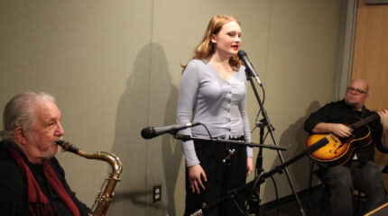 An adult playing tenor saxophone, an adult singing into a microphone, and an adult playing acoustic guitar in a radio studio