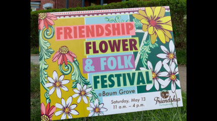 An image of a sign with images of flowers on it posted in grass advertising the Friendship Flower & Folk Festival