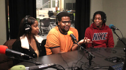 Three teen wearing headphones speaking into microphones in a radio studio