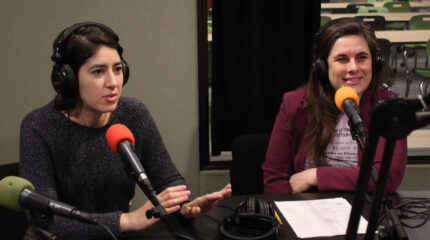 Two adults wearing headphones speaking into microphones in a radio studio