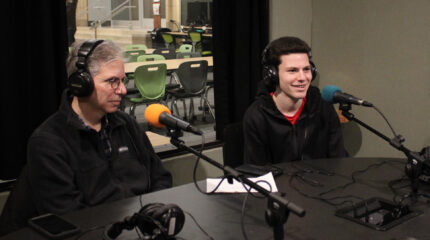 An adult and a youth wearing headphones speaking into microphones in a radio studio