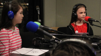 Two youth wearing headphones sitting behind microphones in a radio studio