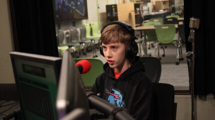 A teen wearing headphones speaking into a microphone in a radio studio
