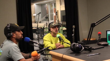 Two adults wearing headphones sitting behind microphones in a radio studio