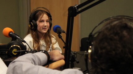 A youth wearing headphones speaking into a microphone being interviewed by an adult wearing headphones in a radio studio