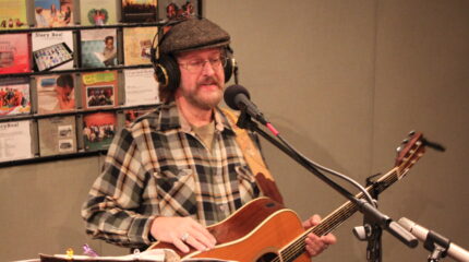 An adult wearing headphones singing into a microphone and playing an acoustic guitar in a radio studio