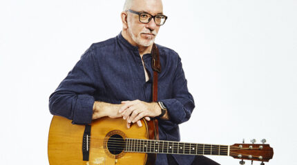 An adult posing and sitting with an acoustic guitar on a white background
